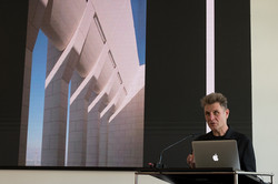 A person gives a lecture to an audience while displaying a presentation slide.