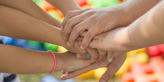 Hände mehrerer Menschen liegen aufeinander. Im Hintergrund sind bunte Sportmatten