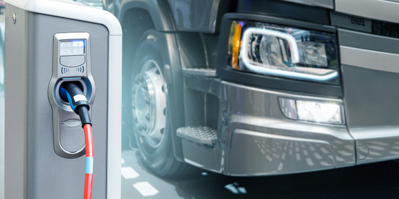 Electric heavy duty transport A gray truck stands next to an electric charging pole with an electric car charging cable plugged into it.