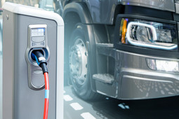 Ein grauer LKW steht neben einer elektrischen Ladesäule, in die ein elektrisches Autoladekabel eingesteckt ist.