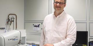 Ein Mann in einem hellen Hemd steht in einem Labor und lächelt in die Kamera.
