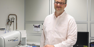 A man in a light-colored shirt stands in a laboratory and smiles at the camera.