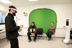 Das Bild zeigt einen "Videostation" in einem weißwändigen Seminarraum. Vorne links im Bild ist eine Person mit einer VR-Brille zu sehen. Zwei auf Stühlen sitzende Personen im Zentrum des Bildes sehen die Person mit VR-Brille an.