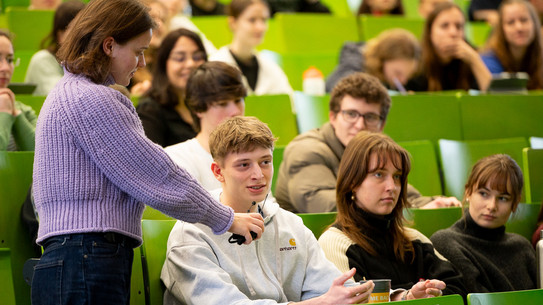 Auf dem Foto sieht man mehrere Personen im Hörsaal sitzen. Im Fokus ist ein junger Mann, der in ein Mikrofron spricht.