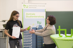 Frau Szymanski übergibt Felix Schlie eine Urkunde und ein Geschenk. Im Hintergrund ist ein TU Banner. Rechts im Bild ein grüner Tisch mit TU-Logo.