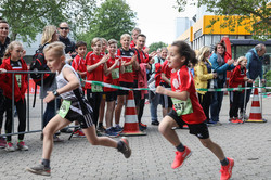 Zwei Kinder rennen beim Kids-Run des Campuslaufs. Im Hintergrund stehen mehrere Personen und feuern die Kinder an.