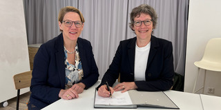 Zwei Frauen mit schwarzen Brillen, kurzen Haaren und schwarzem Blazer sitzen an einem Tisch und lächeln in die Kamera. Die rechte Frau unterschreibt gerade etwas in einer Unterschriftenmappe.