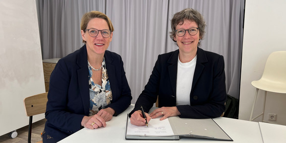 Two women with short hair and who wear glasses and black blazers are sitting at a table and are smiling towards the viewer. The woman on the righthand side is currently signing something in a signature folder.