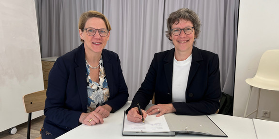Zwei Frauen mit schwarzen Brillen, kurzen Haaren und schwarzem Blazer sitzen an einem Tisch und lächeln in die Kamera. Die rechte Frau unterschreibt gerade etwas in einer Unterschriftenmappe.