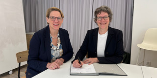 Zwei Frauen mit schwarzen Brillen, kurzen Haaren und schwarzem Blazer sitzen an einem Tisch und lächeln in die Kamera. Die rechte Frau unterschreibt gerade etwas in einer Unterschriftenmappe.
