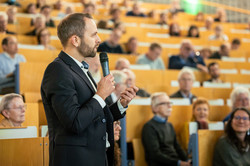 Foto: Eine Person spricht mit einem Mikrofon vor einem Publikum in einem Hörsaal.
