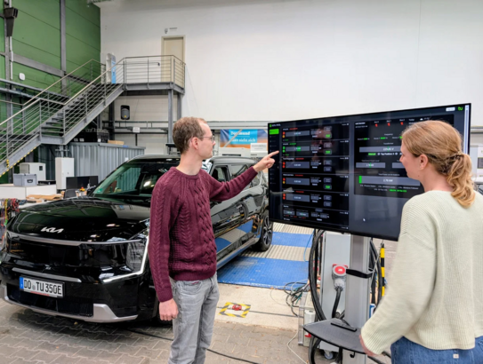 Zwei Personen stehen vor einem Ferseher und analysieren eine Auswertung von dem Auto was im Hintergrund steht.