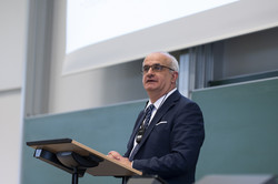 Prof. Bayer, a man in a suit, stands at the lectern in a lecture hall.
