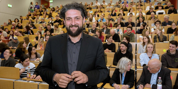 Das Bild zeigt ein Porträtfoto von Prof. El-Mafaalani bei seiner Antrittsvorlesung an der TU Dortmund. Im Hintergrund ist der Hörsaal mit Publikum zu sehen.