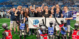 Foto: Die Mitglieder der Forschungsteams Nao Devils (TU Dortmund) und HTWK Robots (HTWK Leipzig) halten Banner in den Händen und recken jubelnd ihre Fäuste in die Luft. Vor den weißen Bannern stehen circa 1,30 Meter große humanoide Roboter in roten und blauen Trikots, die ebenfalls ihre Arme jubelnd in die Luft halten. Die Gruppe steht auf einem grünen Rasenplatz in einem großen Hallenstadion mit gefüllten Rängen.