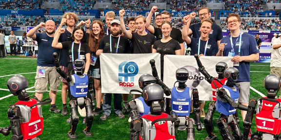 Foto: Die Mitglieder der Forschungsteams Nao Devils (TU Dortmund) und HTWK Robots (HTWK Leipzig) halten Banner in den Händen und recken jubelnd ihre Fäuste in die Luft. Vor den weißen Bannern stehen circa 1,30 Meter große humanoide Roboter in roten und blauen Trikots, die ebenfalls ihre Arme jubelnd in die Luft halten. Die Gruppe steht auf einem grünen Rasenplatz in einem großen Hallenstadion mit gefüllten Rängen.