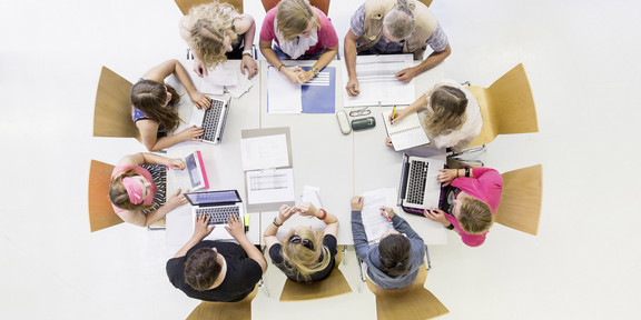 Menschen sitzen mit Arbeitsmarterialien an einem Tisch