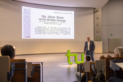 Manfred Bayer steht auf der Bühne vor einer Präsentation über Nobels Initialzündung