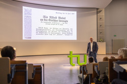 Vortrag Bayer Manfred Bayer steht auf der Bühne vor einer Präsentation über Nobels Initialzündung