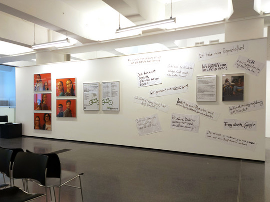 Foto: Eine Ausstellungswand mit orange hinterlegten Porträtfotos, Informationsplakaten und zahlreichen handschriftlichen Notizen, davor eine Stuhlreihe in einem hellen Raum.