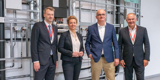 Four people are standing in front of a wall with a technical system consisting of various pipes.