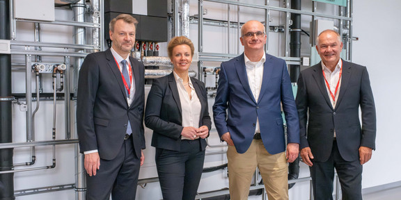 Einweihung TGA-Labor Vier Personen stehen vor einer Wand mit einer technischen Anlage, die aus verschiedenen Rohren besteht.