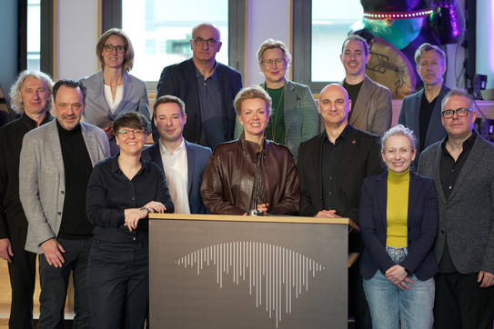 Foto: Eine Gruppe von Personen steht gemeinsam hinter einem Rednerpult bei einer Veranstaltung.