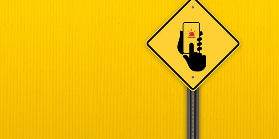 A yellow traffic sign shows two hands holding a cell phone with a flashing warning signal.