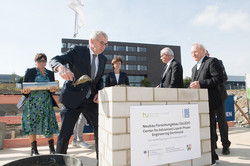 Several people are standing around a brick cube and want to sink a time capsule. A man is filling the cube with a putty knife and putty.