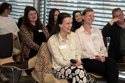 Mehrere Personen sitzen in drei Reihen hintereinander. Frauen in der ersten Reihe lachen.