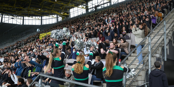 Die mit Erstsemestern besetzte Tribüne. Im Vordergrund Cheerleader, die zu einer LaOla-Welle animieren.