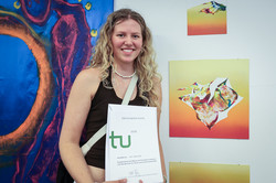 Foto: Eine junge Frau mit blonden lockigen Haaren steht vor einem Kunstwerk und hält eine Urkunde in den Händen.