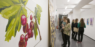 Ausstellung Gartenspäher Ein Gang im Dortmunder U mit Menschen, die sich die Bilder an der Wand ansehen. Links an der Wand hängt ein Bild von gemalten Kirschen im Detail.