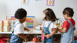 Drei Kleinkinder stehen vor einer niedrigen Ablage mit Spielsachen. Eines der Kinder greift nach einem Stift, der auf der Ablage liegt.