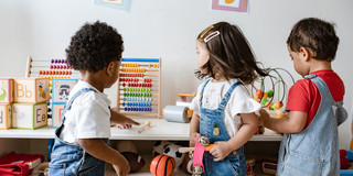 Three toddlers are standing in front of a low shelf with toys. One of the children reaches for a pencil lying on the shelf.