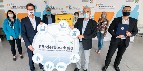 Zu sehen sind mehrere Erwachsene mit Masken. Zwei Männer halten ein Plakat hoch. Im Hintergrund sind Banner mit der Aufschrift Ruhr-Konferenz zu sehen.