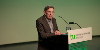 A gentleman with glasses and a suit stands at a lectern labeled "TU Dortmund".