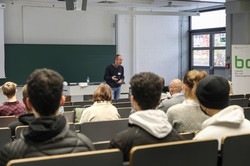 Several people are sitting in a seminar building, a man is speaking in front of a green board.