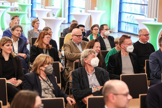 Das Publikum sitzt in Reihen, im Hintergrund ist eine Fensterfront, vor der Tische stehen.