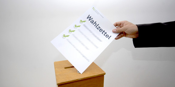 Bild einer beliebigen Wahlurne, in die ein Zettel eingeworfen wird.