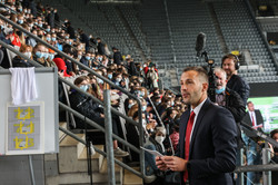 A man in a suit stands in a soccer stadium in the stands full of first-year students and is filmed and photographed by two members of the press.