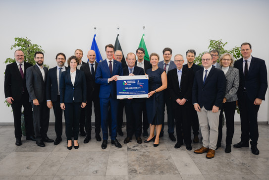 Foto: Vertreter*innen des Projekts präsentieren gemeinsam den Förderbescheid vor den Flaggen von Europa, Deutschland und Nordrhein-Westfalen.