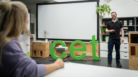 Mann präsentiert vor Leinwand in einem Raum mit großen grünen „cet“-Buchstaben, während eine Person im Vordergrund zuhört.