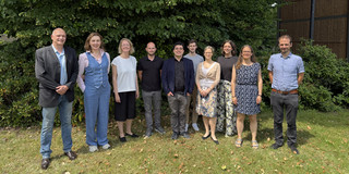 Ten people are standing side by side for a group photo. There is a green tree in the background.