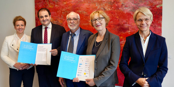 Ein Gruppenfoto von 5 Personen vor einem rot-orangenem Gemälde. Zwei Personen halten blaue Mappen.