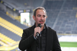 Ein Mann hält ein Mikrofon in der Hand. Im Hintergrund die leeren Tribünen des BVB-Stadions.