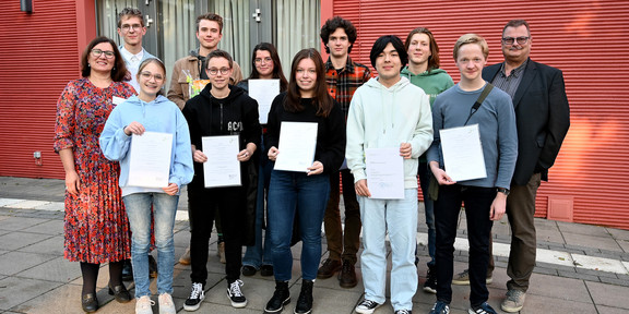 Neun Jugendliche halten Urkunden in den Händen und posieren für ein Gruppenfoto. Drei Erwachsene stehen links und rechts von der Gruppe.