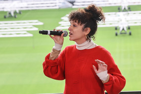 Eine Sängerin steht in einem Fußballstadion, hält ein Mikrofon in der Hand und singt.