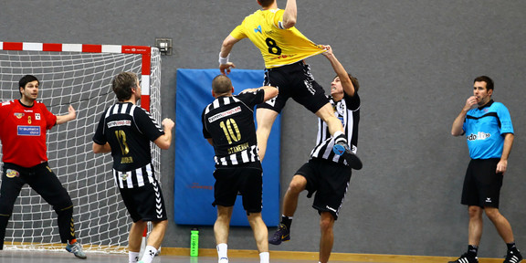 A handball player jumps in the air to throw a goal, players of the opposing team try to stop him from doing so