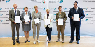 Four women and two men stand with documents in their hands in front of a display wall with the inscription "Bildungsland NRW".